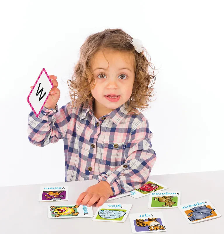 A young child with curly hair sits at a table holding up an Orchard Toys Alphabet Flashcard featuring the letter "W." The child, dressed in a plaid shirt, is surrounded by various colorful animal-themed cards from the set scattered on the table.