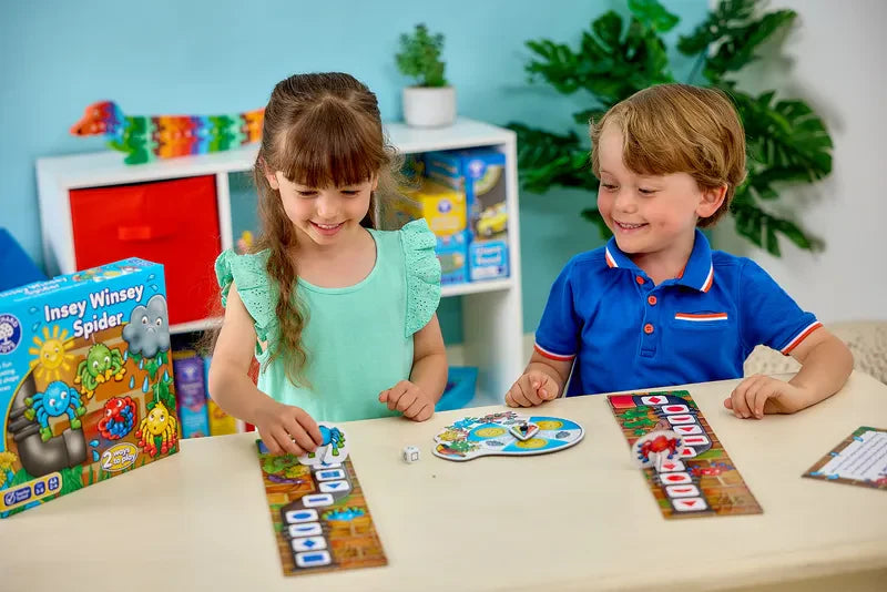 Two children are playing a board game called "Orchard Toys Insey, Winsey Spider" at a table. The girl is moving a piece on her board, while the boy smiles beside her. The room has colorful shelves and a potted plant in the background.