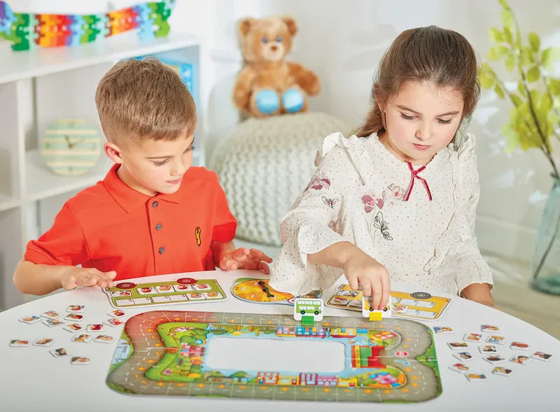 Two children are engaged in a game of Orchard Toys Bus Stop at a table. The boy, dressed in a red shirt, is focusing intently on his pieces, while the girl, wearing a white dress, places a yellow piece onto the board. In the background, a stuffed teddy bear can be seen.