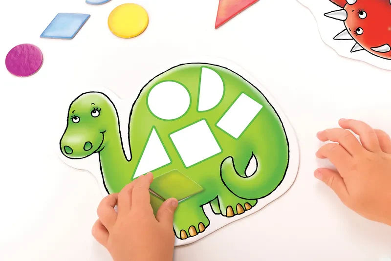 A child's hands place vibrant geometric shapes into the Orchard Toys Dotty Dinosaurs puzzle, featuring a smiling green dinosaur with cutouts. The background is white, with various shapes like circles, squares, and triangles scattered around.