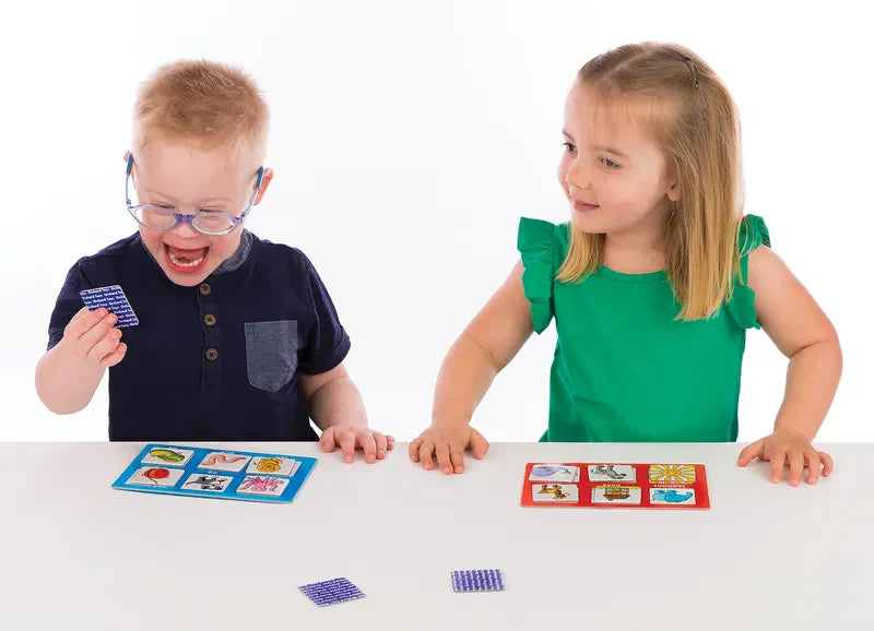 A boy with glasses and Down syndrome, dressed in a navy shirt, eagerly holds an Orchard Toys Alphabet Lotto card while sitting next to a girl in a green dress. They are playing the matching card game together at a white table.