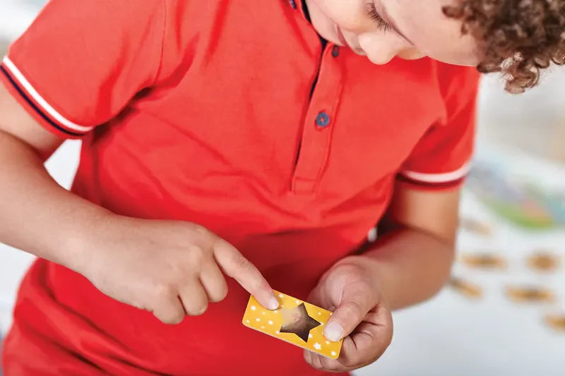 A child in a red shirt is examining the Orchard Toys Magic Maths card, which is yellow and features a star-shaped cutout. They are pointing at the card that's held in their other hand, with the background appearing blurred.