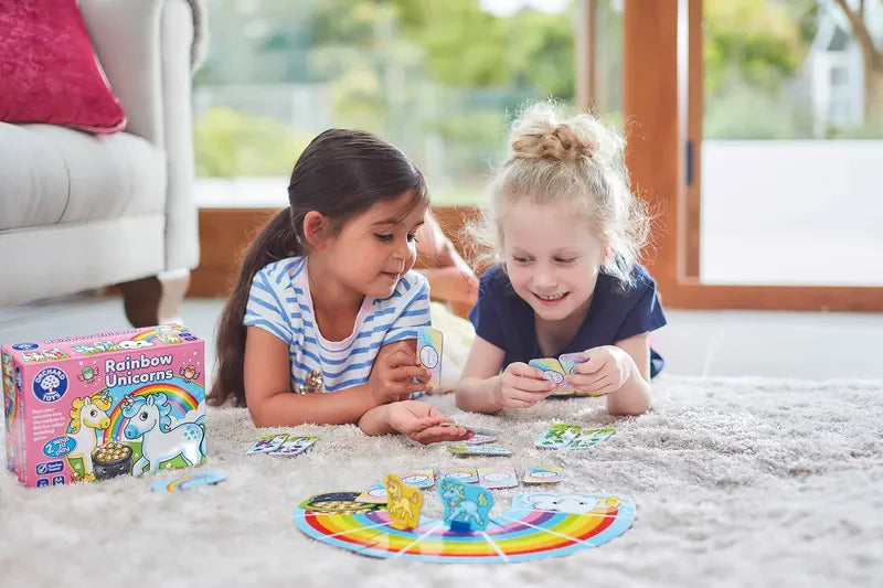 Two young girls are lying on a soft rug, playing the vibrant "Orchard Toys Rainbow Unicorns" board game. They're engaged and smiling, surrounded by cards and game pieces as sunlight streams in through a large window in the background.