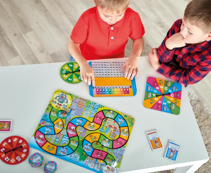 Two children sit at a white table playing Orchard Toys Times Tables Heroes. One child in a red shirt looks at the number board, while the other in a red and black shirt observes. The table holds game pieces, cards, and a spinner.