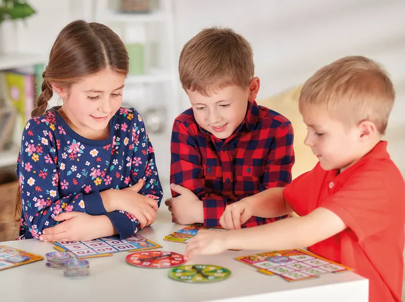 Three children are playing with the "Orchard Toys Times Tables Heroes" game at a table. A girl wearing a floral shirt and two boys, one in a plaid shirt and the other in a red shirt, are absorbed in the game pieces and cards before them. The room is bright and cheerful.