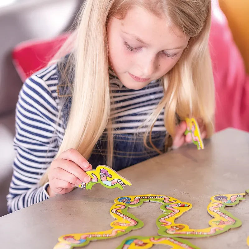 A young girl with long blonde hair and a striped shirt is focused on putting together the Orchard Toys Wiggly Words puzzle with its colorful, whimsical pieces on a table. She holds two pieces in her hands, concentrating on fitting them together.