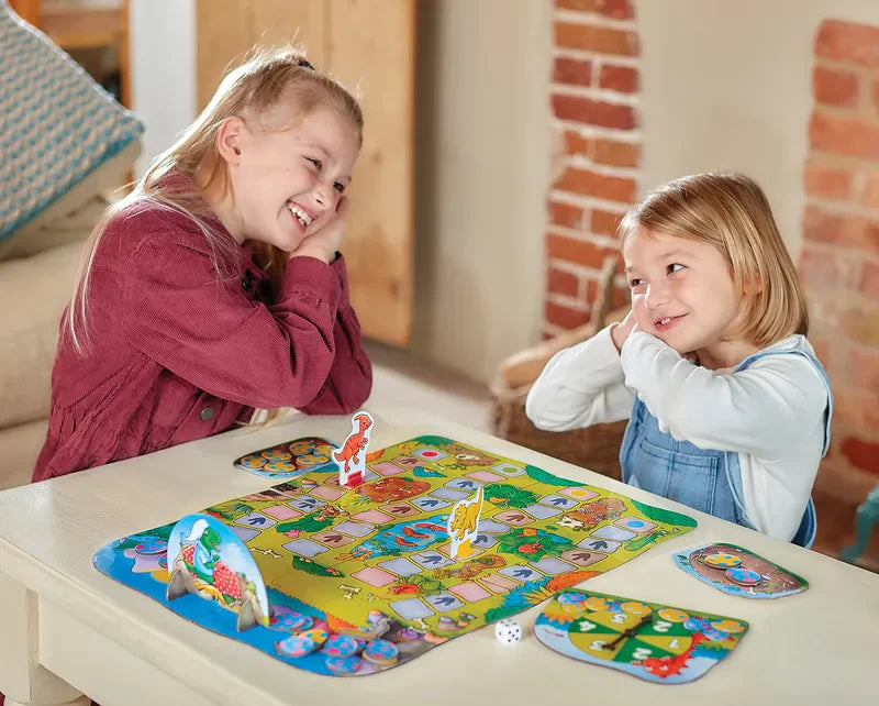 Two children sit at a table, smiling with their hands on their cheeks, as they enjoy playing the colorful Orchard Toys Dino-Snore-Us board game. The room has brick walls and a cozy atmosphere.