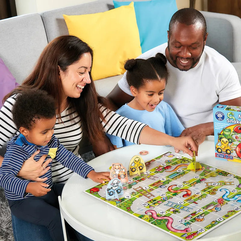 A family of four enjoys playing "Orchard Toys My First Snakes & Ladders." They are seated around a small table, smiling and looking engaged. The toddler is held by the mother, while the older child moves a piece on the board. A game box is visible.