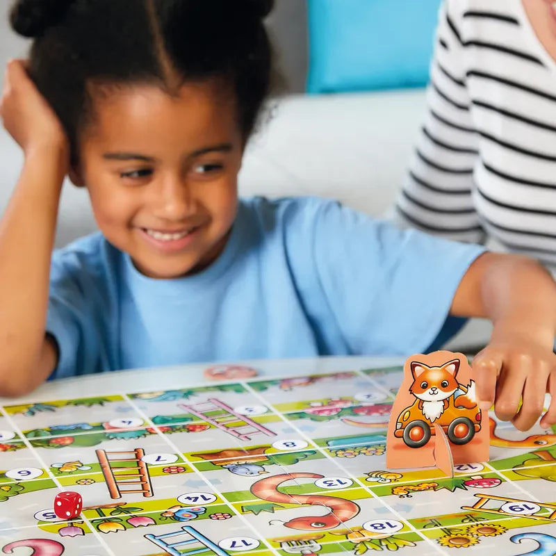 A child grins while enjoying the Orchard Toys My First Snakes & Ladders board game, which features vibrant snakes, ladders, and a fox-shaped game piece. The board is vividly illustrated as another person sits nearby, partially in view.