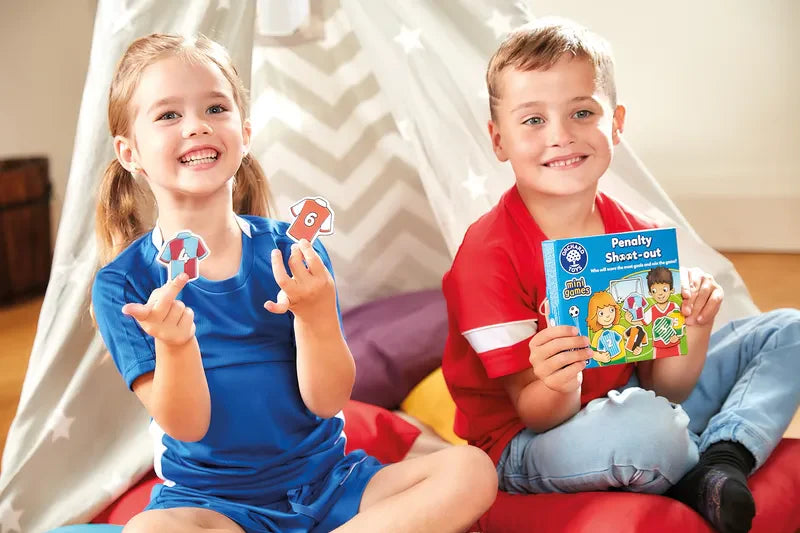 Two smiling children sit in front of a play tent. The girl, dressed in a blue outfit, is holding game pieces, while the boy, in a red shirt, holds the box of an Orchard Toys Penalty Shoot Out Mini Game. Colorful cushions surround them.