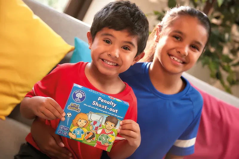 Two children are sitting on a couch, smiling. The child on the left, wearing a red shirt, holds the Orchard Toys Penalty Shoot Out Mini Game. The child on the right is wearing a blue shirt. A plant can be seen in the background.