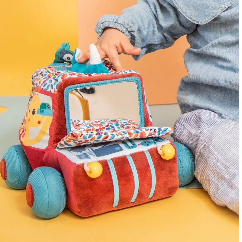 A child wearing a light blue outfit explores a colorful Lilliputiens I'm Discovering Activities Car Safari. The soft toy truck features blue wheels, a floral roof, a reflective window, and stuffed animal figures on top with various fabric textures and patterns.