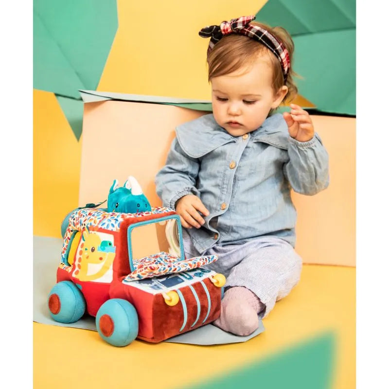 A toddler in a denim shirt and grey pants sits on a colorful background, exploring sensory activities. With a headband bow, the child gazes at the Lilliputiens I'm Discovering Activities Car Safari toy vehicle with a small stuffed dinosaur, captivated by this delightful baby activity.