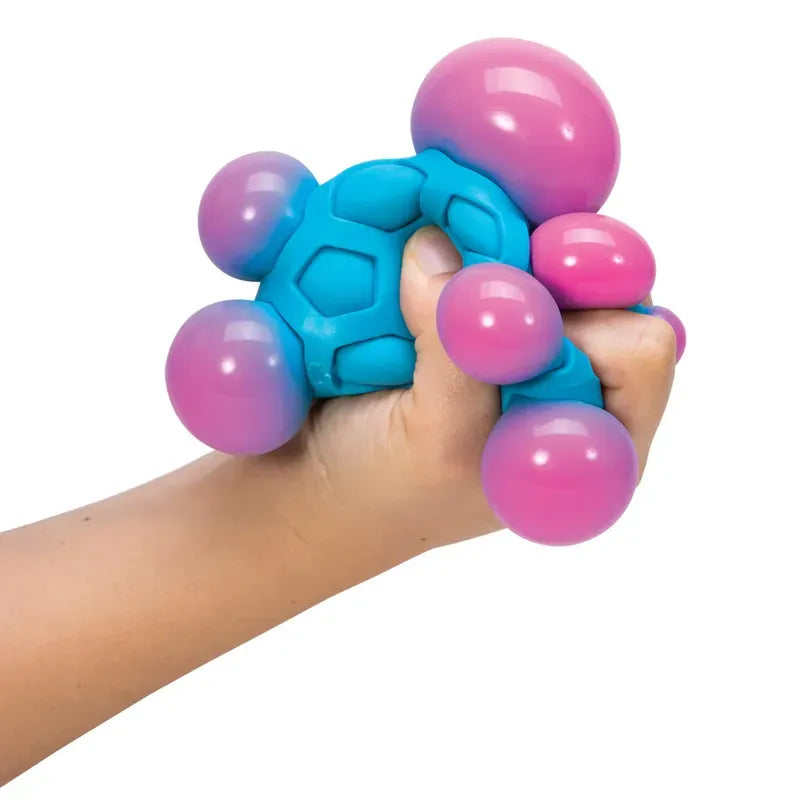A hand grips the Atomic Needoh, a blue stress ball with pink and purple orbs. This fidget toy boasts a flexible design and textured surface for enhanced tactile satisfaction.