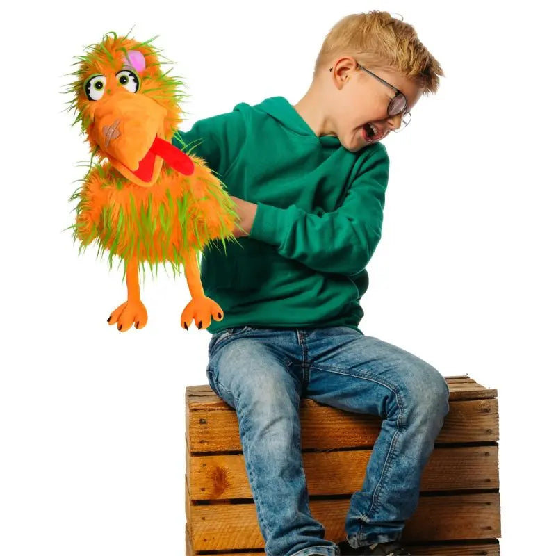 A smiling boy in glasses and a green hoodie plays with the Living Puppets Bernie the Bird Hand Puppet, an orange and green character, while sitting on a wooden crate against a white background.