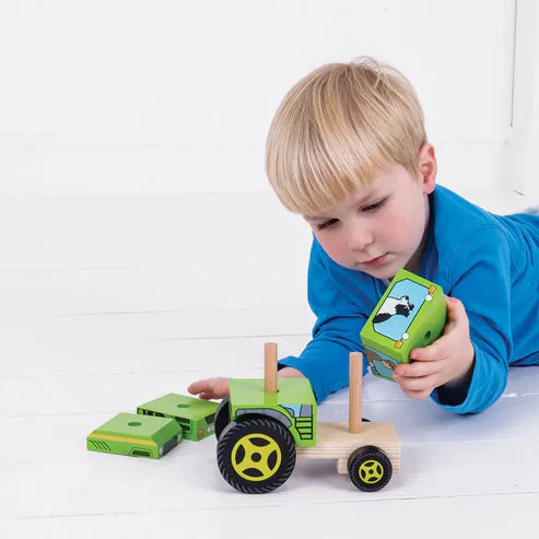 A young blond boy in a blue shirt lies on a white floor, playing with the Bigjigs Stacking Tractor and its detachable parts, developing fine motor skills as he explores and assembles each piece.