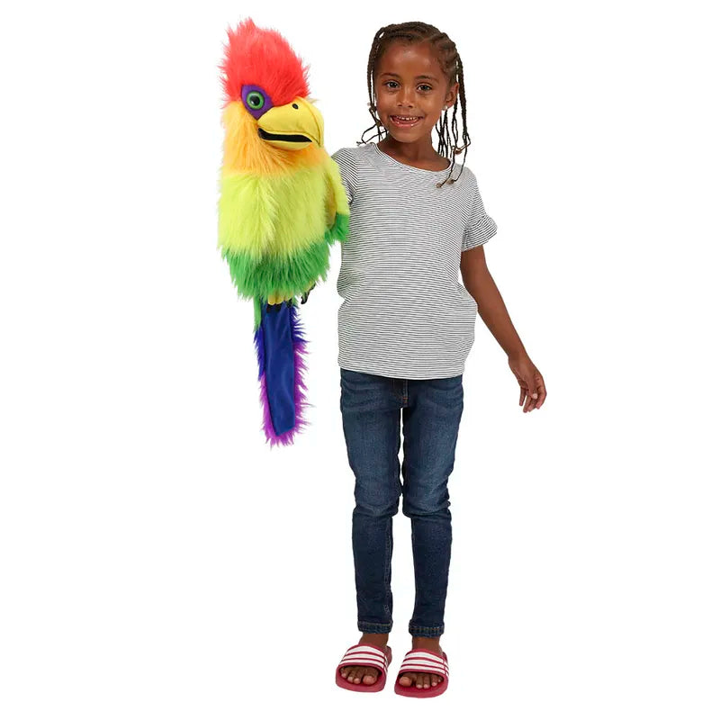 A young girl with braided hair, dressed in a striped gray t-shirt, blue jeans, and red sandals is holding a stunning Puppet Company Rainbow Bird Hand Puppet. The vibrant puppet features fuzzy feathers and a yellow beak with a squeaker in the mouth, extending from her hand.