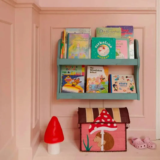 A pastel pink nursery wall features a teal bookshelf with colorful books, the Lamp Small Mushroom Red, a handcrafted lamp, a toy chest with hedgehog and mushroom designs, and pink clogs—a charming touch for children's room decor.
