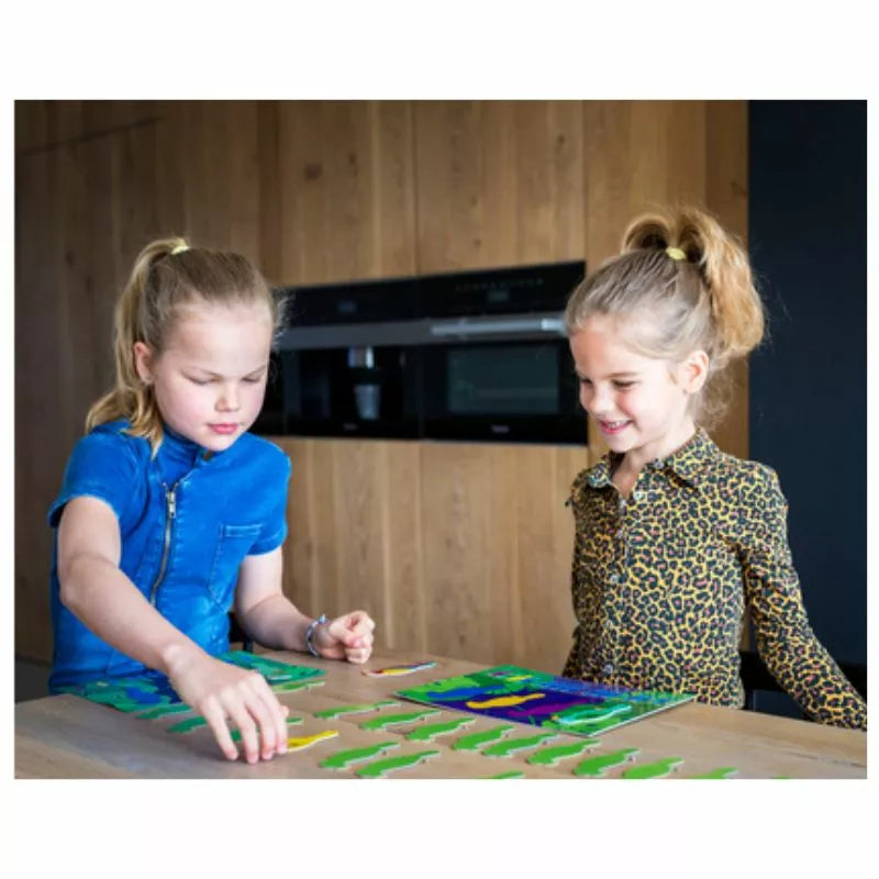 Two children with ponytails are captivated by the Buitenspeel Parrots Collection Game at a wooden table. They concentrate on arranging green pieces, surrounded by colorful parrots adorning the wooden wall and black appliances in the background. One child is dressed in blue, while the other sports a leopard print outfit as they plan their next moves.