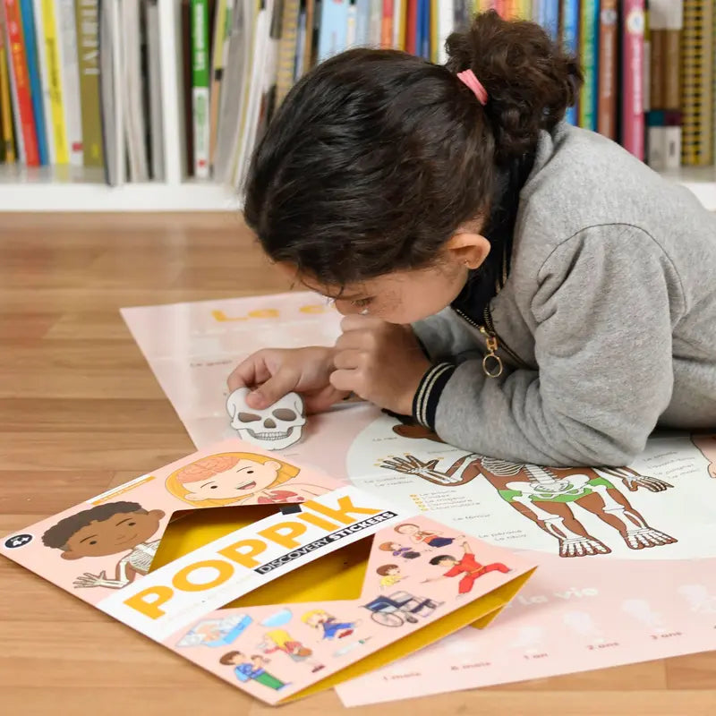 A young child places a paper skull on a colorful Poppik Discovery Sticker Poster – Human Body, with the Poppik educational stickers booklet open nearby and books visible on shelves in the background.