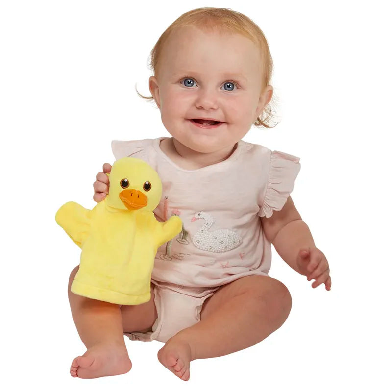 A smiling baby in a light pink outfit with a swan design plays interactively with The Puppet Company My First Puppet Duck, a yellow hand puppet. The blue-eyed, light-haired baby sits against a white background, exploring early language development.