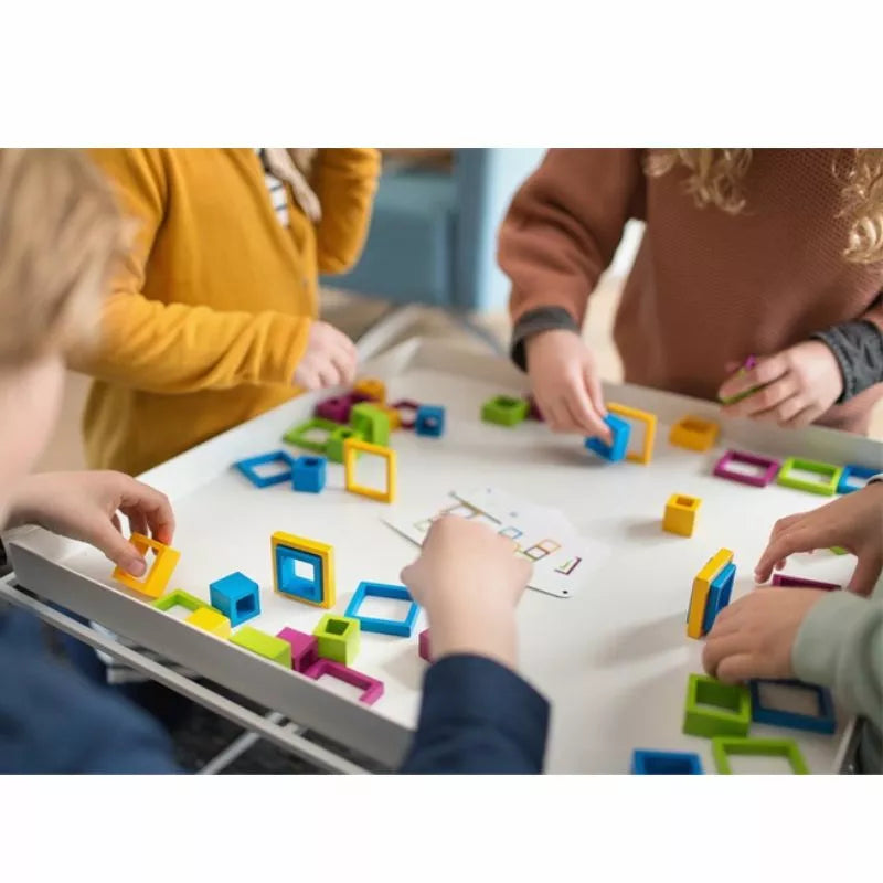 Children's hands are arranging pieces from the Buitenspeel Frames Game on a white table, engaging in a skillful activity that promotes collaborative play. The game features colorful geometric blocks, including squares and rectangles, encouraging quick thinking as kids delve into building patterns and exploring the intriguing world of shapes.