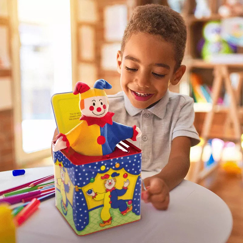 A young boy with a joyful expression plays with a colorful Jester Jack In Box toy, featuring sturdy construction, in a brightly lit room filled with art supplies.