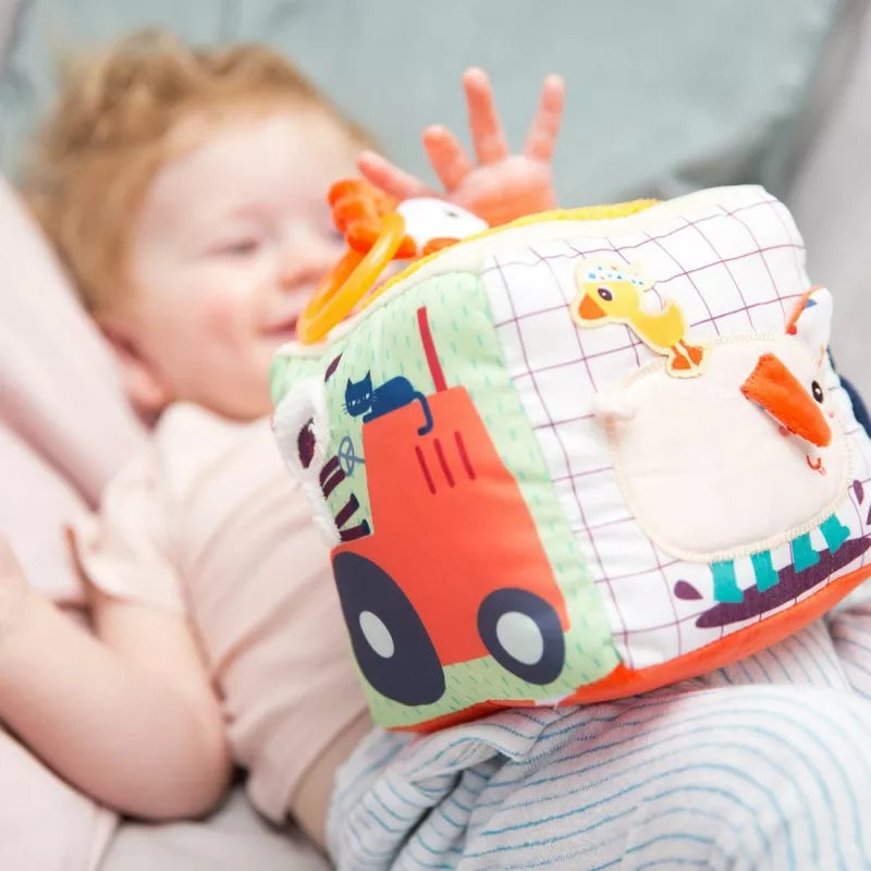 A baby laying on top of a Lilliputiens Farm Activity Sound Cube next to a pillow.