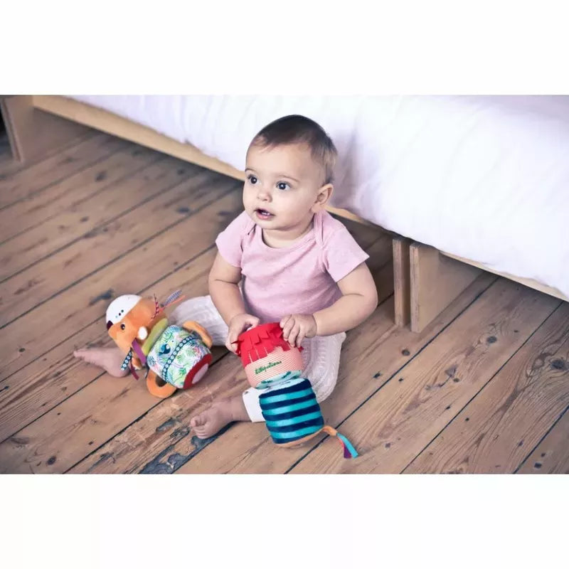 A baby sitting on a wooden floor playing with a Lilliputiens Jack Sound Roll toy, including a toy lion, next to a white couch.