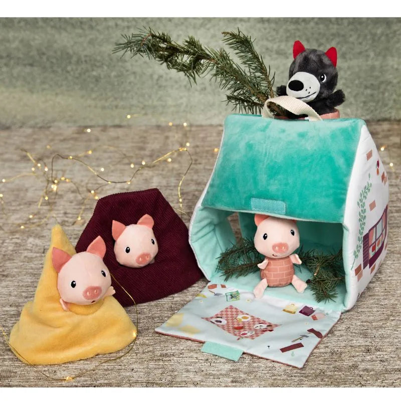 The "Lilliputiens Houses Of The Wolf And The 3 Little Pigs" plush toys encourage creative play by featuring two pig toys wrapped in felt blankets, another pig situated inside a cozy fabric toy house, and a wolf toy peering over the house. Fairy lights and branches add to the enchanting scene. It's perfect for fostering fine motor skills!