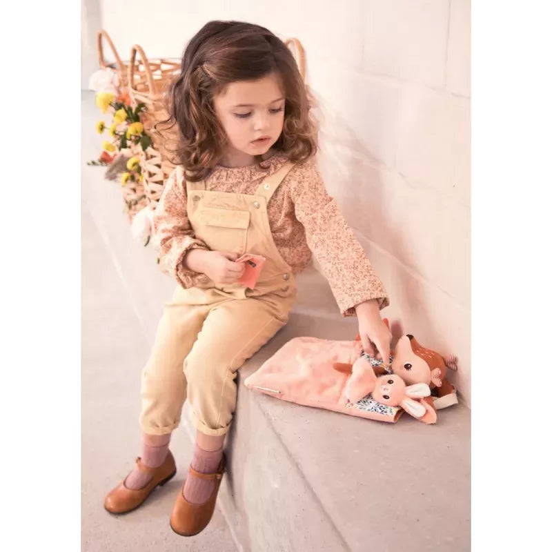 A little girl with curly hair, dressed in a floral shirt and overalls, sits on a step playing with a Lilliputiens Stella Bed Time Ritual toy on a soft pink blanket, ready for bedtime.