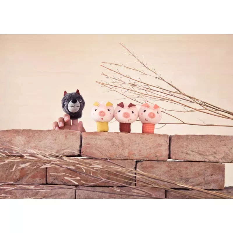 Four whimsical Lilliputiens Louis The Wolf And The 3 Little Pigs Finger Puppets perched on a rustic brick ledge, with delicate dried grasses adding a touch of nature's elegance in the background.