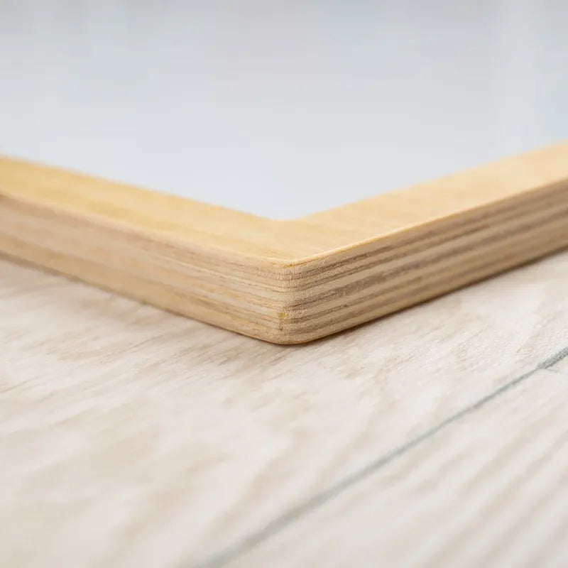 Close-up of the corner of the Bigjigs Magnetic Board on a wooden surface, ideal for showcasing creativity. Its light wood finish with visible grain contrasts sharply with the slightly textured surface beneath.