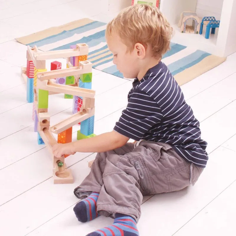 A child joyfully plays on a white wooden floor with the Bigjigs Marble Run, an educational toy. The young one intently guides a marble along the colorful track, while a striped rug and more toys are seen in the background.