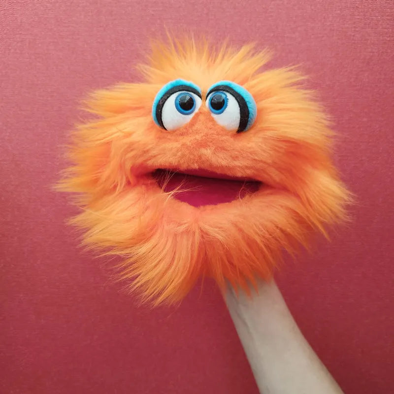 A fuzzy, bright orange hand puppet named Living Puppets Klops, featuring blue googly eyes and a wide-open mouth, is shown against a pink background. The mouth-moving puppet appears to be animatedly expressing itself, held up by a person's hand—perfect for creative play sessions.