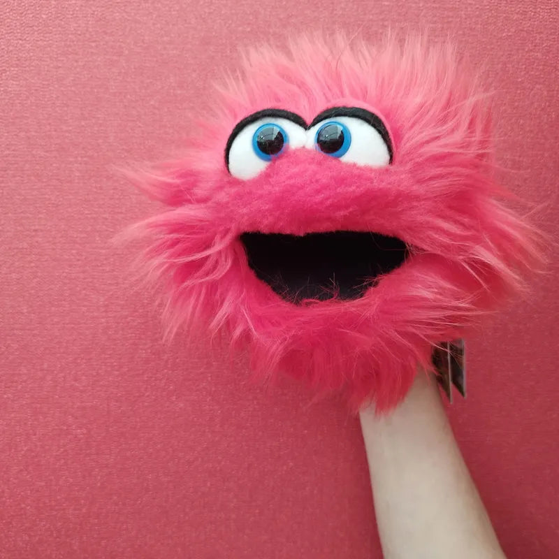 A person holds up the Living Puppets Twaddle Hand Puppet, showcasing its wide eyes and open mouth against a pink background. This fluffy hand puppet exudes a friendly and playful expression, making it perfect for imaginative play.