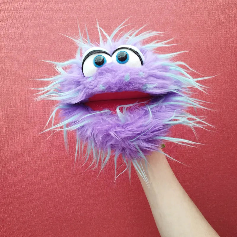 A person's hand holds up the fluffy, shaggy-furred Living Puppets Monster Hand Puppet Tusch with its big round eyes and a red, movable mouth against a pink background. Featuring playful blue accents, this whimsical purple puppet exudes charm and fun.