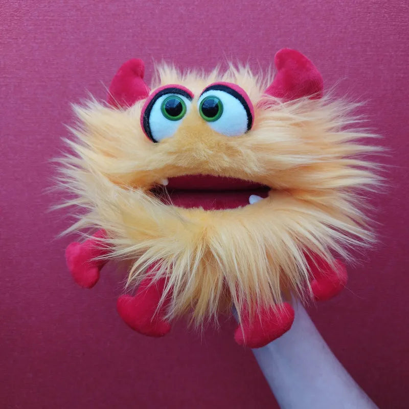 A person holds the Living Puppets Monster Hand Puppet Gisa, which resembles a furry yellow and red monster with bright green eyes, a wide open mouth, and small red horns and limbs. The solid deep pink background makes the puppet pop vividly.