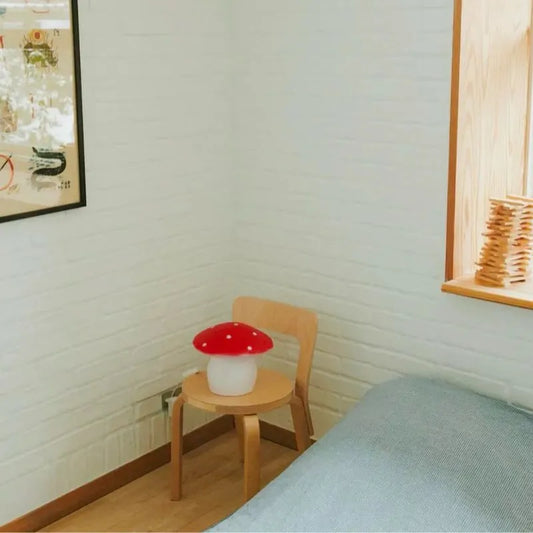 A cozy, minimal bedroom corner features a wooden chair and the Lamp Medium Mushroom Red, set beside a bed with a grey blanket, a window ledge, and a framed picture on a white brick wall.