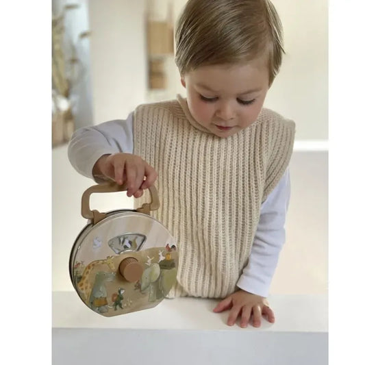 A young child in a cream knit vest and white shirt looks down while holding the Musical Radio Noah's Ark, a small toy suitcase adorned with animal illustrations.