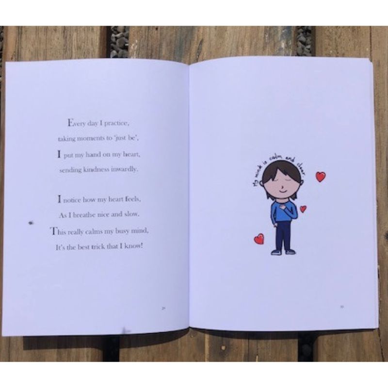 An open book lays on a wooden surface. The left page contains a poem about practicing mindfulness. On the right, an illustration from *My Busy Popcorn Mind* shows a smiling person with short hair, wearing a blue shirt and holding their heart, surrounded by three small red hearts reflecting self-compassion.