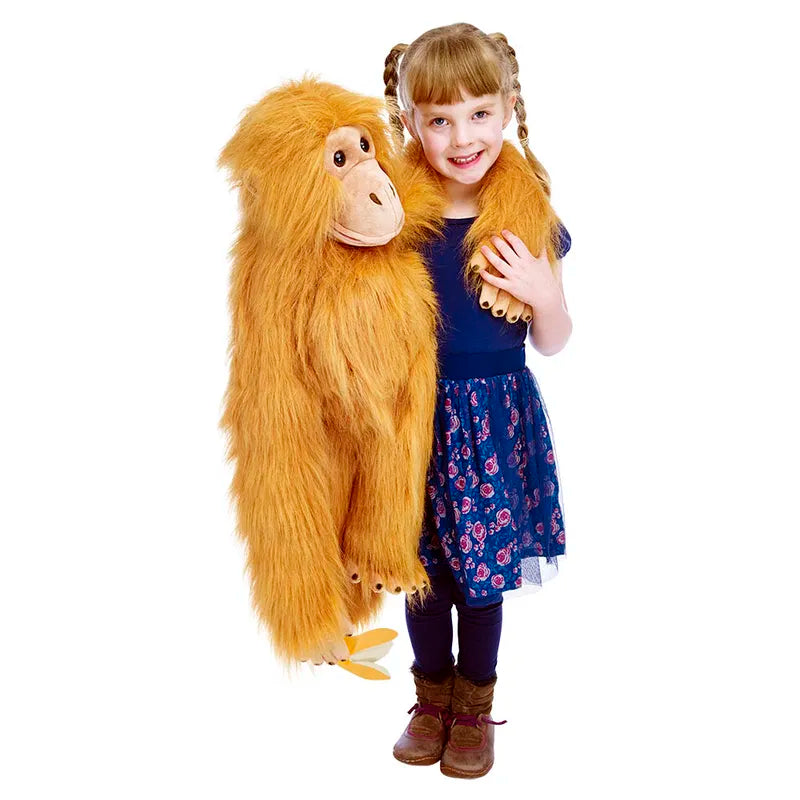 A smiling young girl with pigtails and a floral skirt holds "The Puppet Company Large Puppet Orangutan." She is wearing a blue top and brown boots. The orangutan puppet, featuring vibrant orange fur and a friendly expression, appears almost lifelike as she skillfully moves its mouth like an experienced puppeteer.