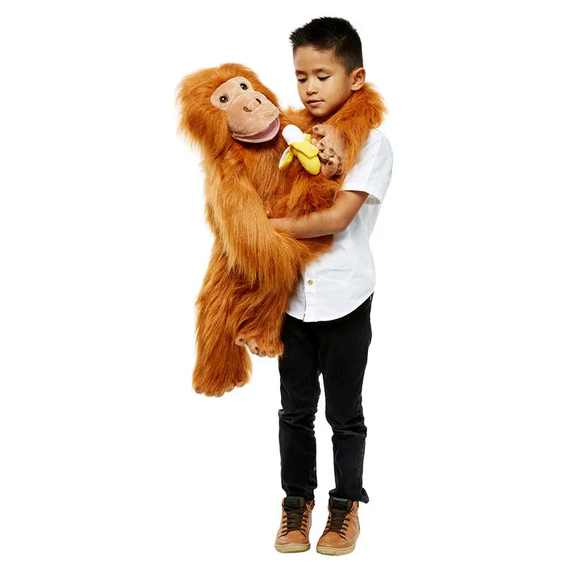 A boy stands with The Puppet Company Large Puppet Orangutan. This vibrant puppet, with its bright orange fur and big smile, features hands that can move its mouth. It clutches a small banana as the boy, dressed in a white shirt and dark pants, gazes at it with a neutral expression.