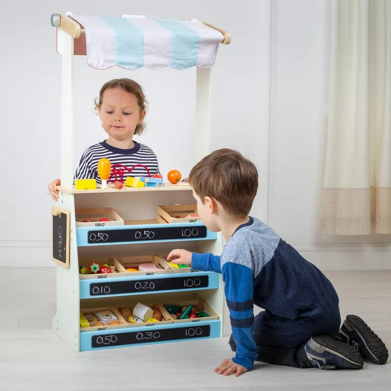 Two young children play with the Tidlo Playshop and Puppet Theatre with FREE Finger Puppets. The girl stands behind the striped canopy, while the boy kneels in front, choosing items from labeled shelves for imaginative role-play.