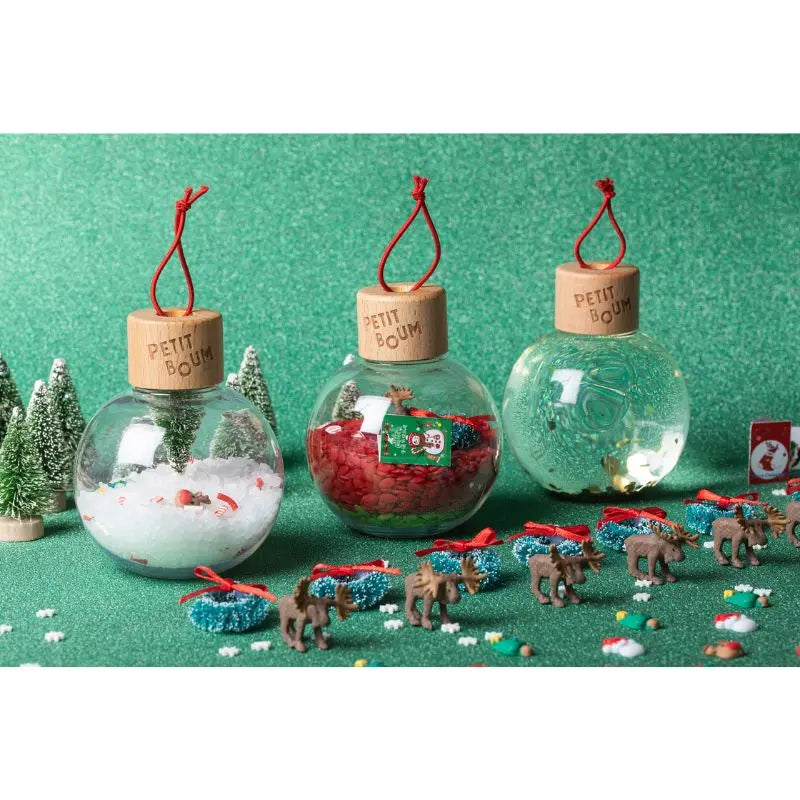 Three handmade Christmas Sensory Baubles from Spain—clear globes with wooden lids, red strings, snow, red beads, and water—rest on a green surface among mini decorated trees and festive ornaments.