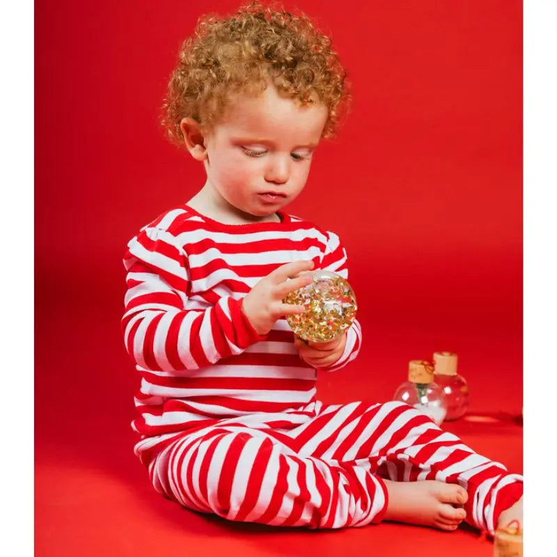 A young child in red and white striped pajamas sits on the floor against a red background, gazing at a sparkling Christmas Sensory Bauble handmade in Spain.