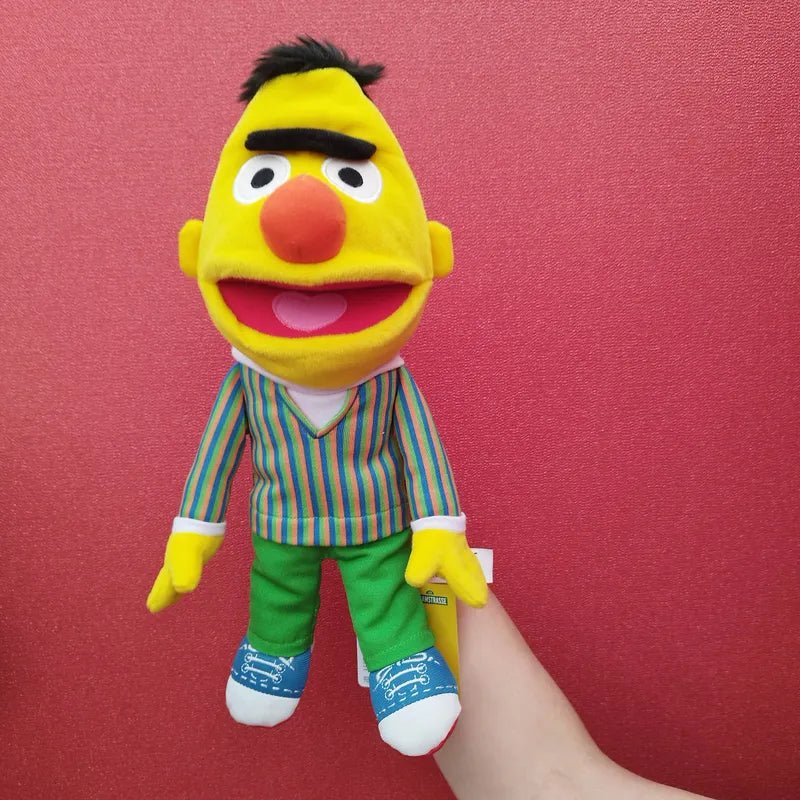 A person holds a Living Puppets Bert Hand Puppet 33cm from Sesame Street against a red background. This Sesame Street puppet, with its yellow face, orange nose, black hair with a unibrow, and dressed in a striped shirt, green pants, and blue sneakers is not only adorable but also easy to use for children and adults.