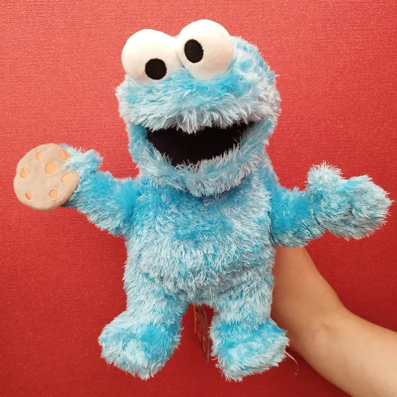 A hand is holding the Living Puppets Cookie Monster Hand Puppet, a blue, fluffy Sesame Street character with large googly eyes and a wide, open mouth. The toy clutches a small fabric cookie in one hand, perfect for storytelling and social skills, set against a bright red background.