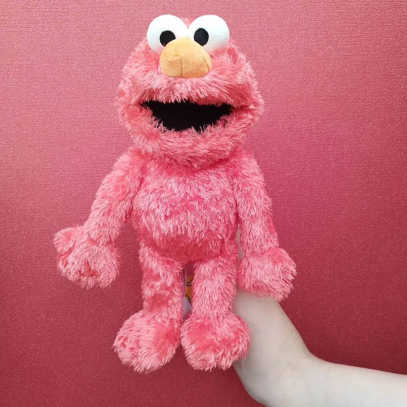 The Living Puppets Elmo Sesame Street Hand Puppet, a pink plush storytelling toy with big eyes, an orange nose, and an open mouth giving it a cheerful appearance, is held up against a pink background by a hand visible in the lower right corner of the image.