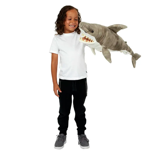 A child with long hair in a white T-shirt and black pants smiles while holding "The Puppet Company Large Creature Shark" plush toy. Their imagination soars as they engage in creative play, gazing at the open-mouthed shark puppet against a plain white backdrop.